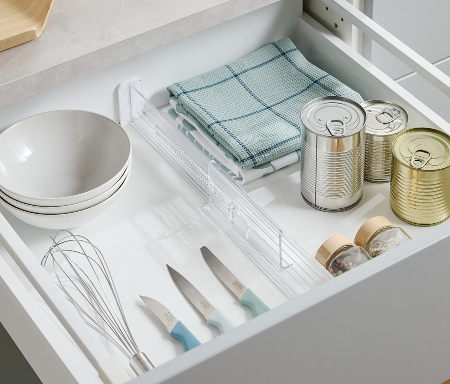 Pohľad zhora na otvorenú zásuvku s miskami, utierkou, plechovkami, korením, nožmi a metličkou.