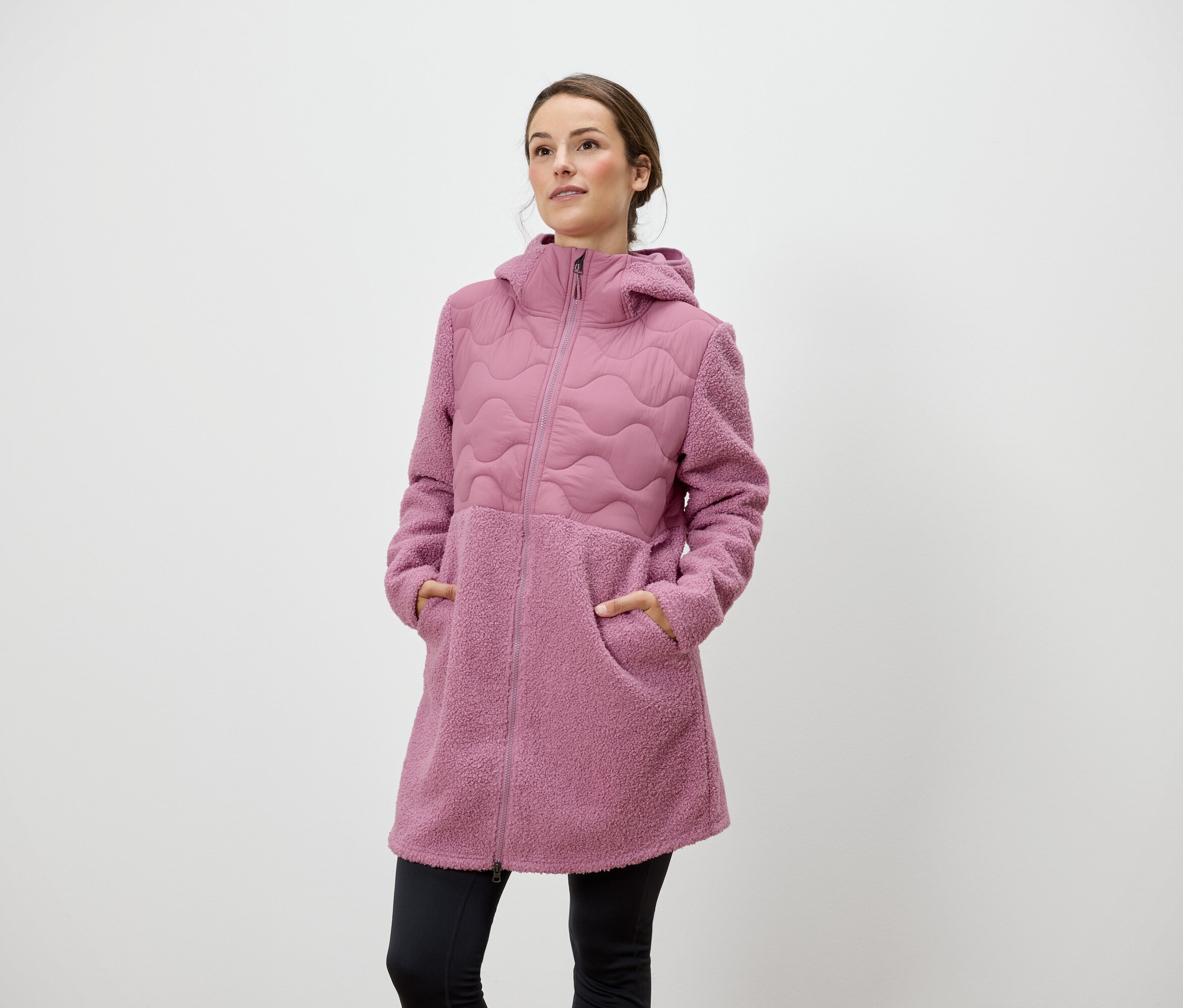 Žena stojí a má ruky vo vreckách ružového kabáta so zipsom a kapucňou.