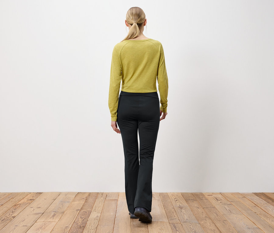 Žena s blond vlasmi stiahnutými do copu stojí chrbtom k fotoaparátu a má na sebe olivovozelený top s dlhým rukávom a čierne nohavice.