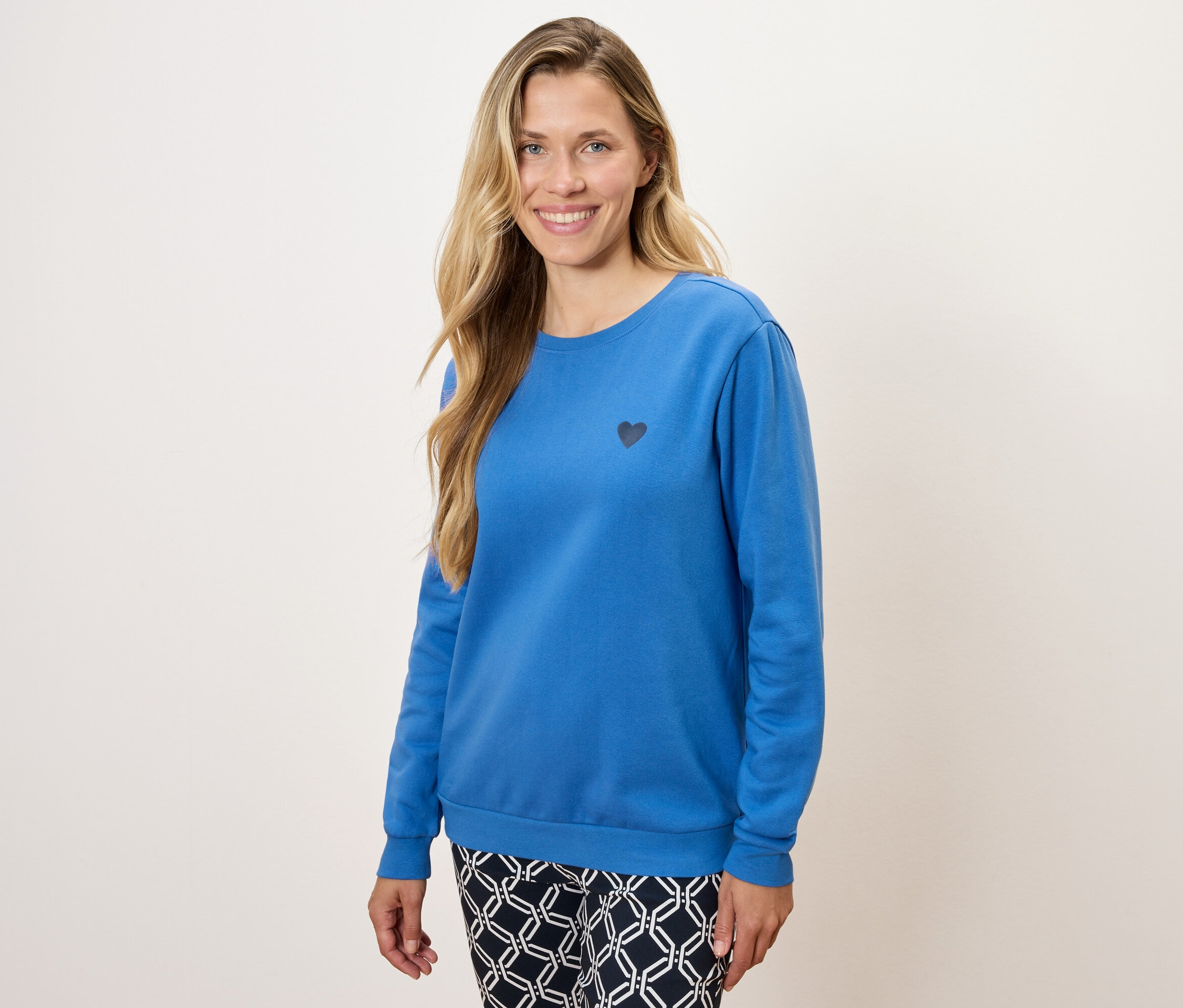 Žena má na sebe modrú mikinu s dlhým rukávom s malým čiernym srdcom a vzorované nohavice.