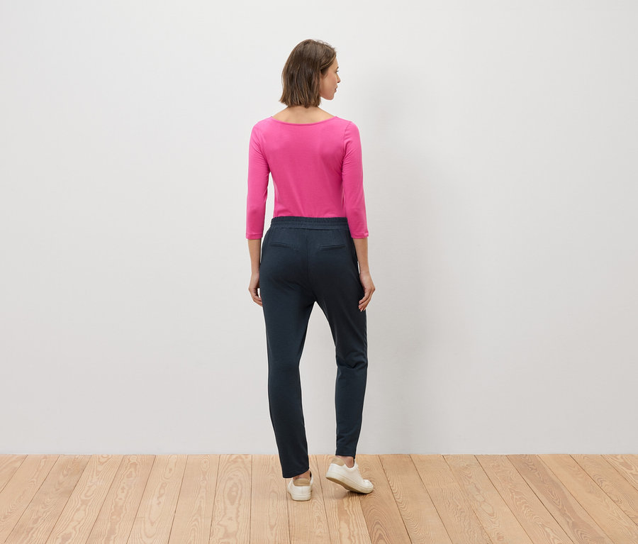 Žena stojí chrbtom k fotoaparátu, má na sebe ružovú blúzku a modré nohavice.