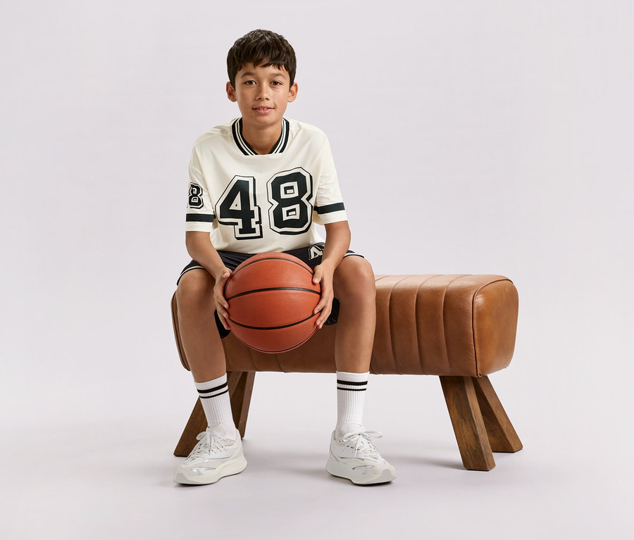 Chlapec sedí na hnedej lavičke a drží basketbalovú loptu.