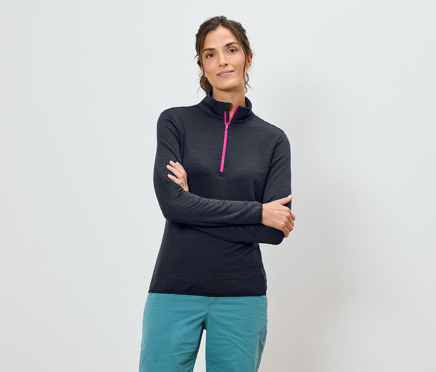 Žena so skríženými rukami má na sebe tmavomodrú košeľu s dlhým rukávom s ružovým zipsom a tyrkysové nohavice na bielom pozadí.