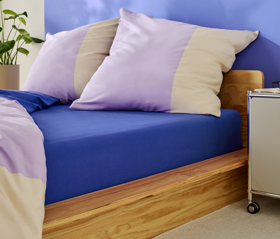 Posteľ s modrou napínacou plachtou a vankúšmi s fialovými a béžovými pruhmi.