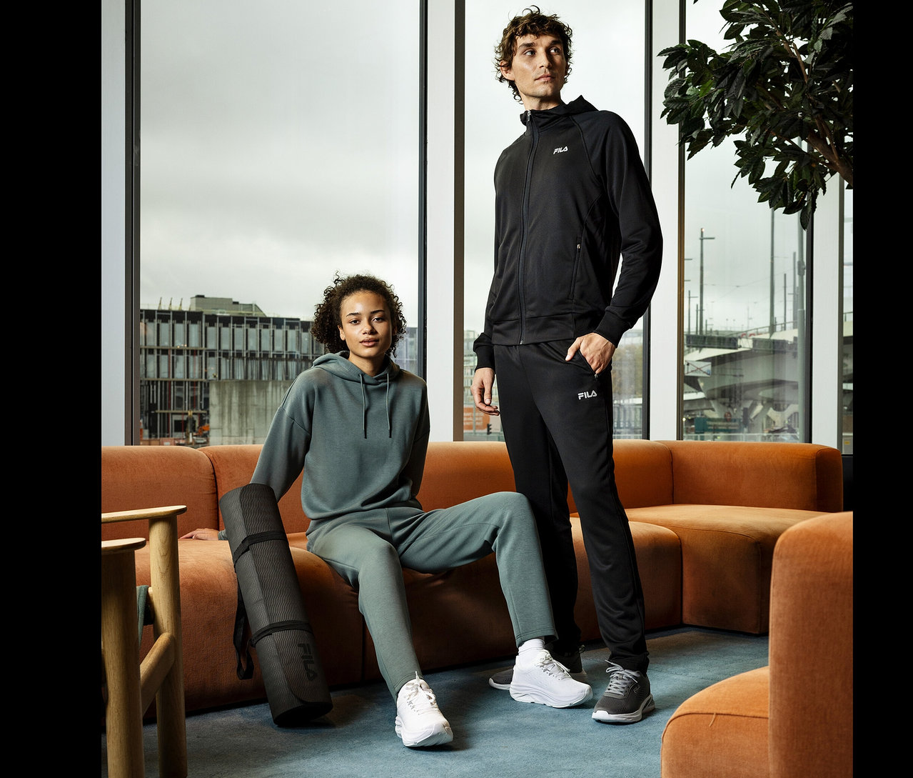 Žena sedí na oranžovej pohovke s čiernou podložkou na jogu. Vedľa nej stojí muž. Obaja majú na sebe športové oblečenie značky Fila.