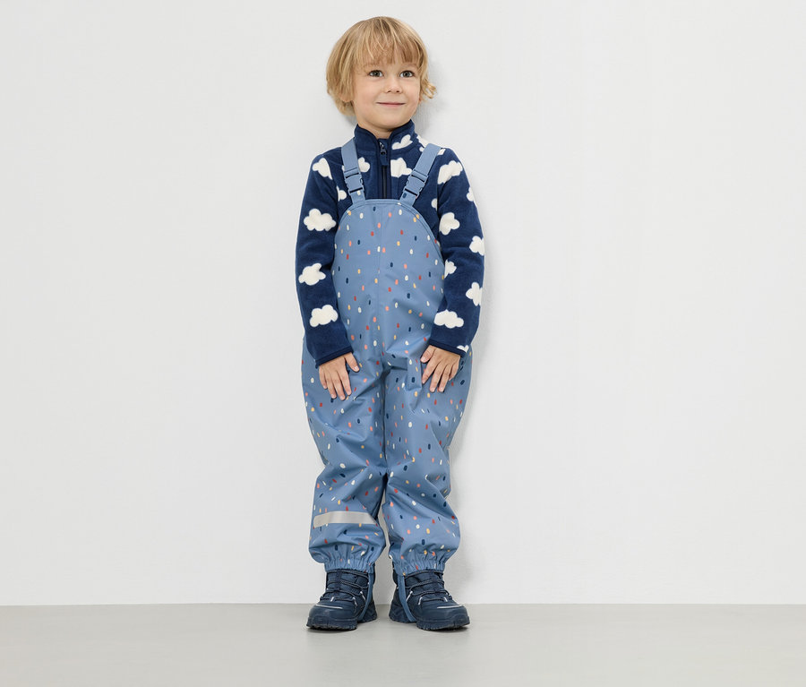 Malý chlapec stojí pred bielou stenou a má na sebe modrú fleecovú bundu s obláčikovým vzorom a modré dažďové nohavice s farebnými kvapkami.