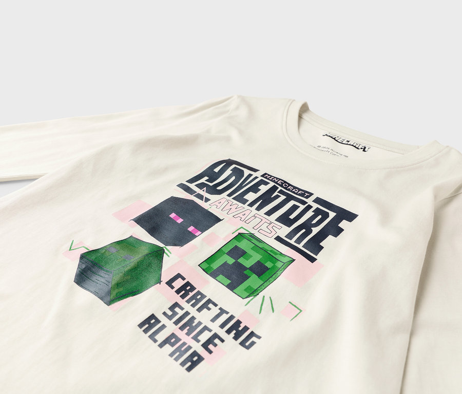 Na bielom tričku s dlhým rukávom je dizajn Minecraft s nápisom "Adventure Awaits" a "Crafting Since Alpha".