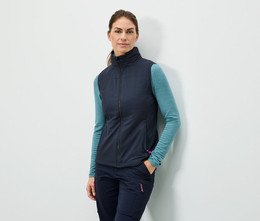 Žena stojí s rukou vo vrecku pri stene, má na sebe tmavomodrú vestu, tyrkysové tričko s dlhým rukávom a tmavomodré nohavice.