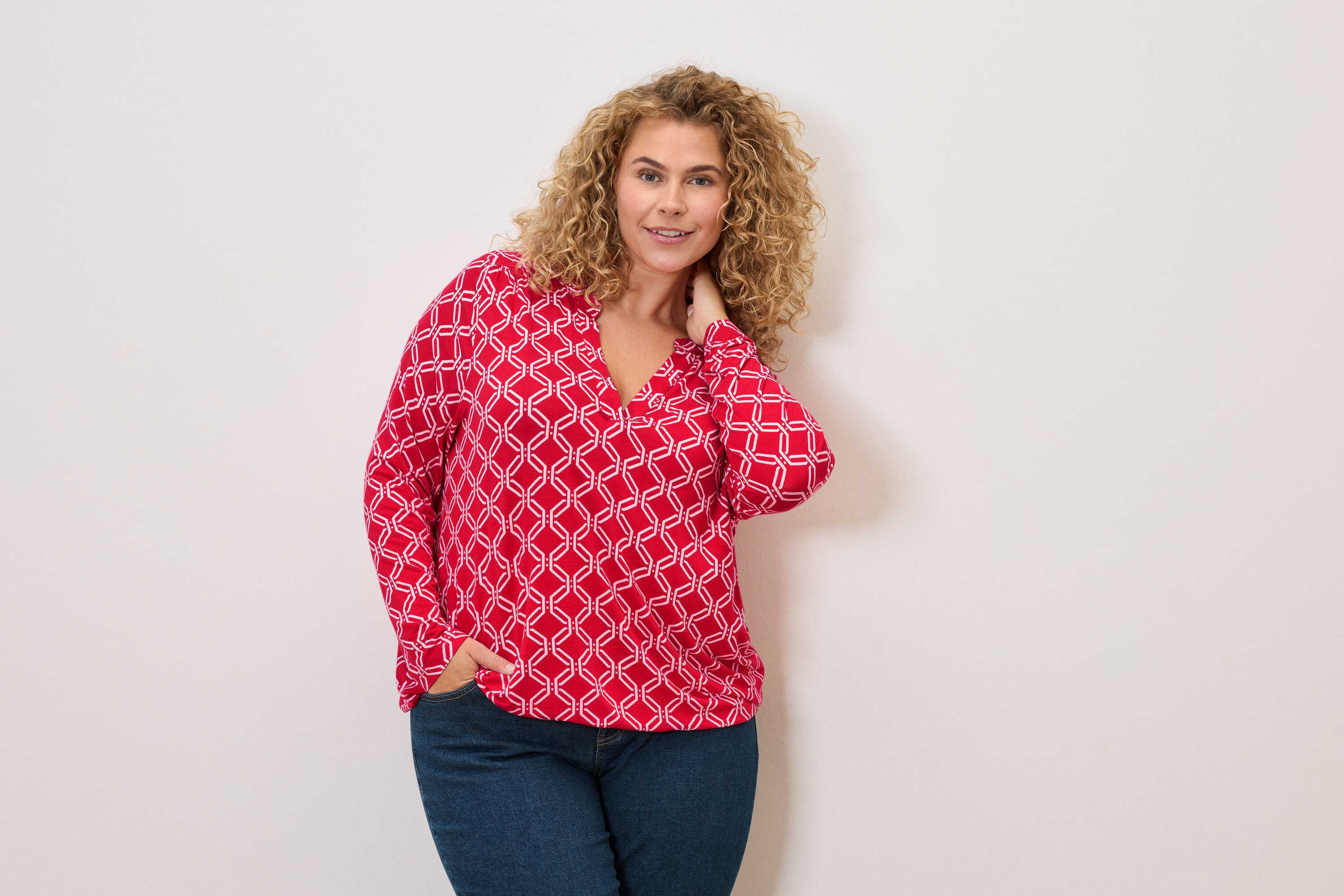 Žena s kučeravými vlasmi v červeno-bielej blúzke a džínsoch pózuje s rukou vo vrecku.