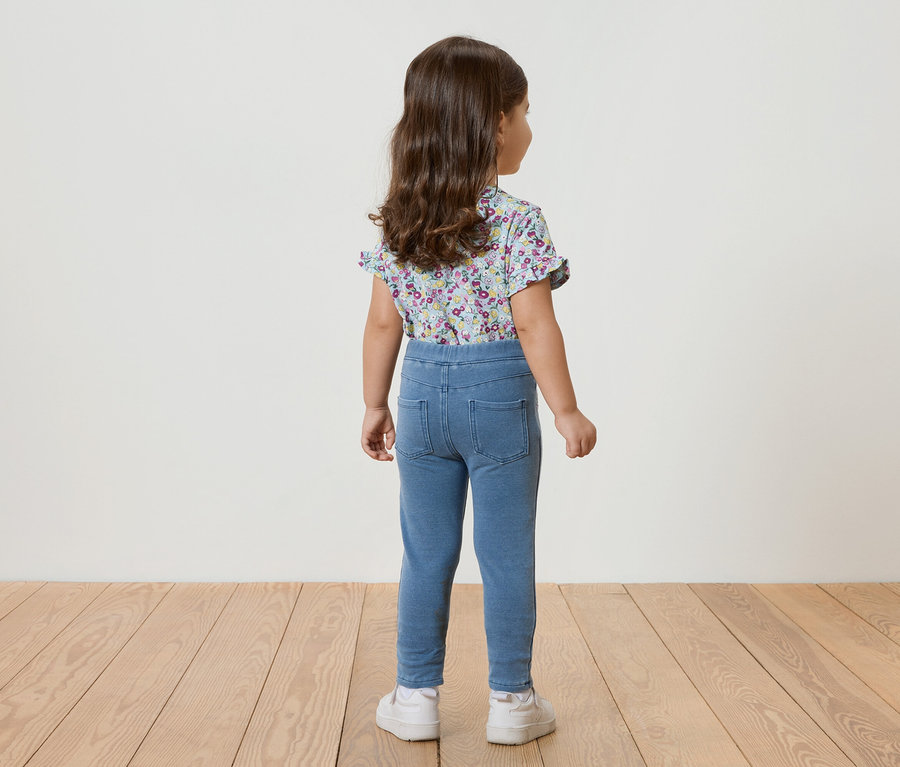 Pohľad zozadu na malé dievča s kučeravými vlasmi v modrom kvetinovom tope s krátkym rukávom, modrých legínach a bielych teniskách.