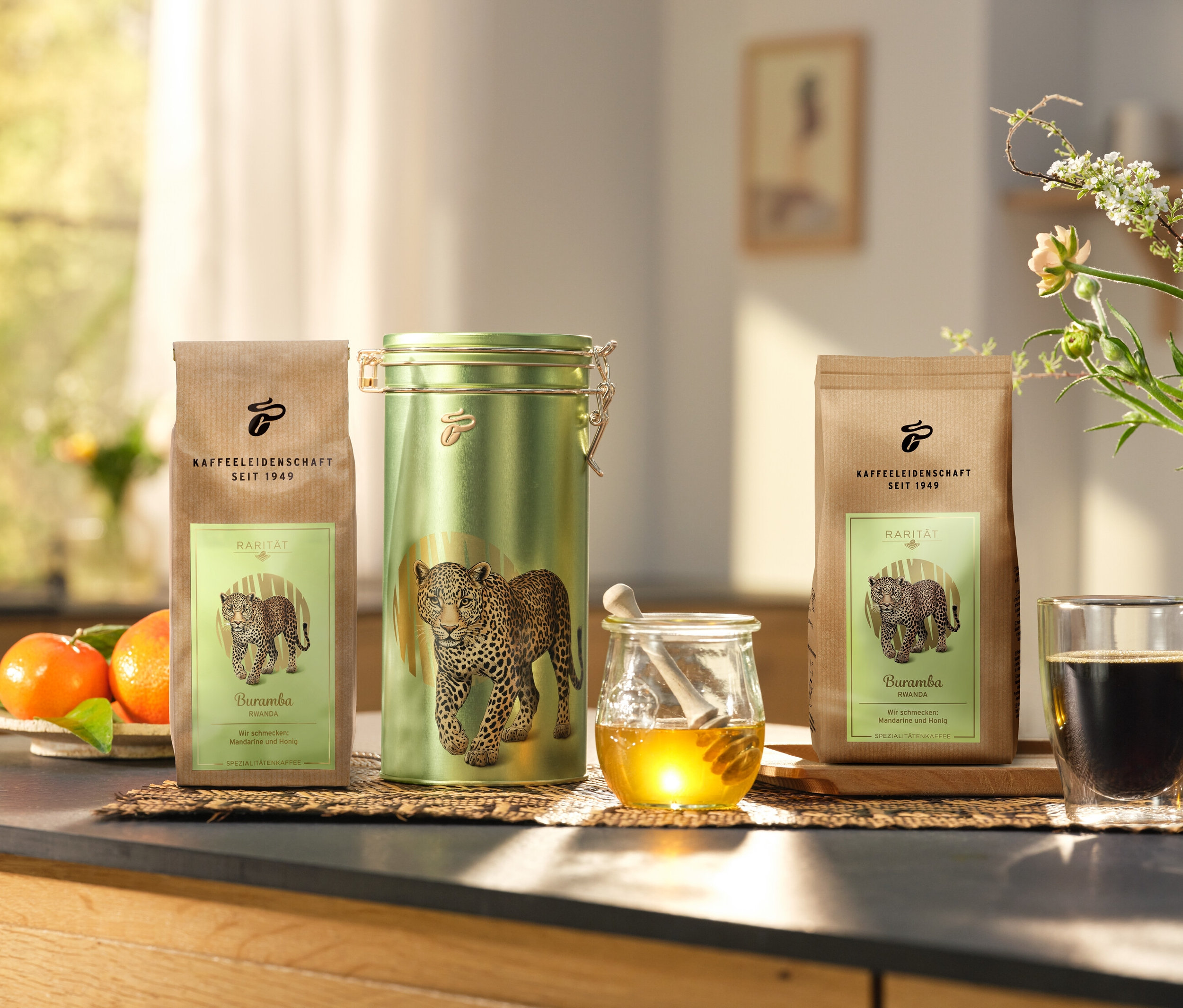 Na kuchynskej linke stojí dóza na kávu a dve balenia »Buramba Rwanda« vrátane dózy na kávu vedľa šálky kávy a medu.