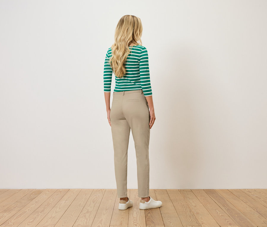 Žena s blond, vlnitými vlasmi stojí chrbtom k divákovi, oblečená v zeleno-bielom pruhovanom tope, béžových nohaviciach a bielych teniskách.