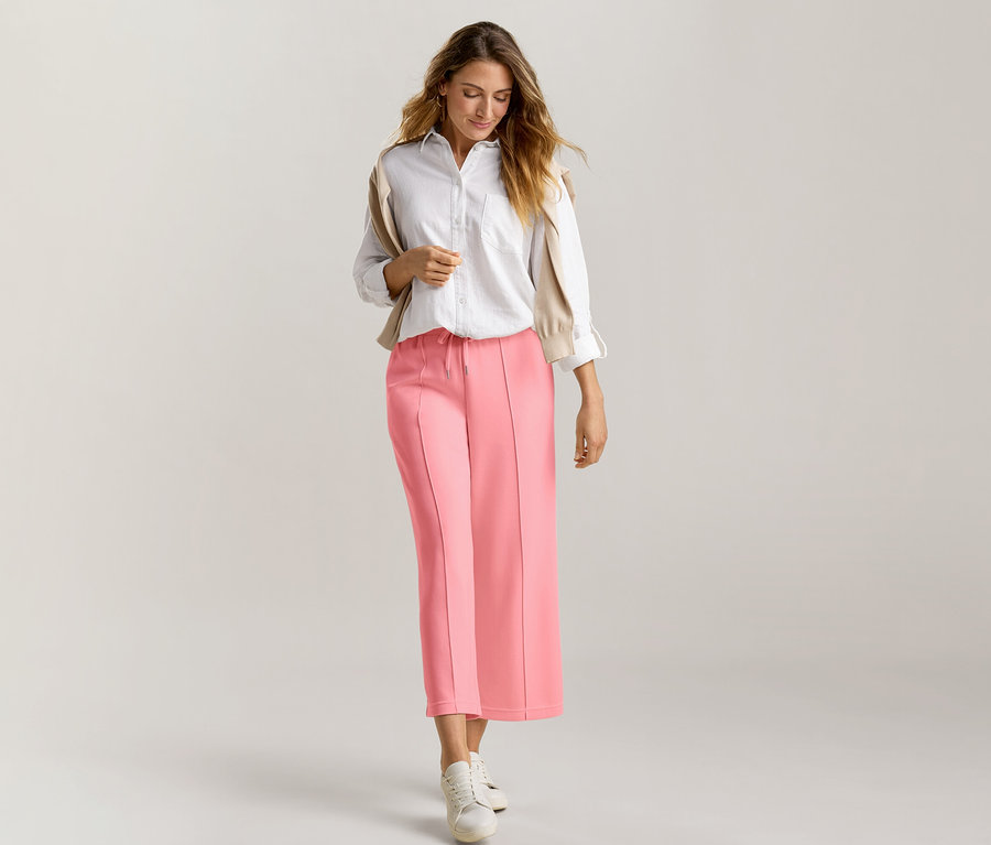 Žena s dlhými hnedými vlasmi kráča dopredu a pozerá sa dole. Má na sebe bielu košeľu, béžový sveter prehodený cez plecia a koralovo ružové nohavice culotte zo zmesi modalu, skombinované s bielymi teniskami.