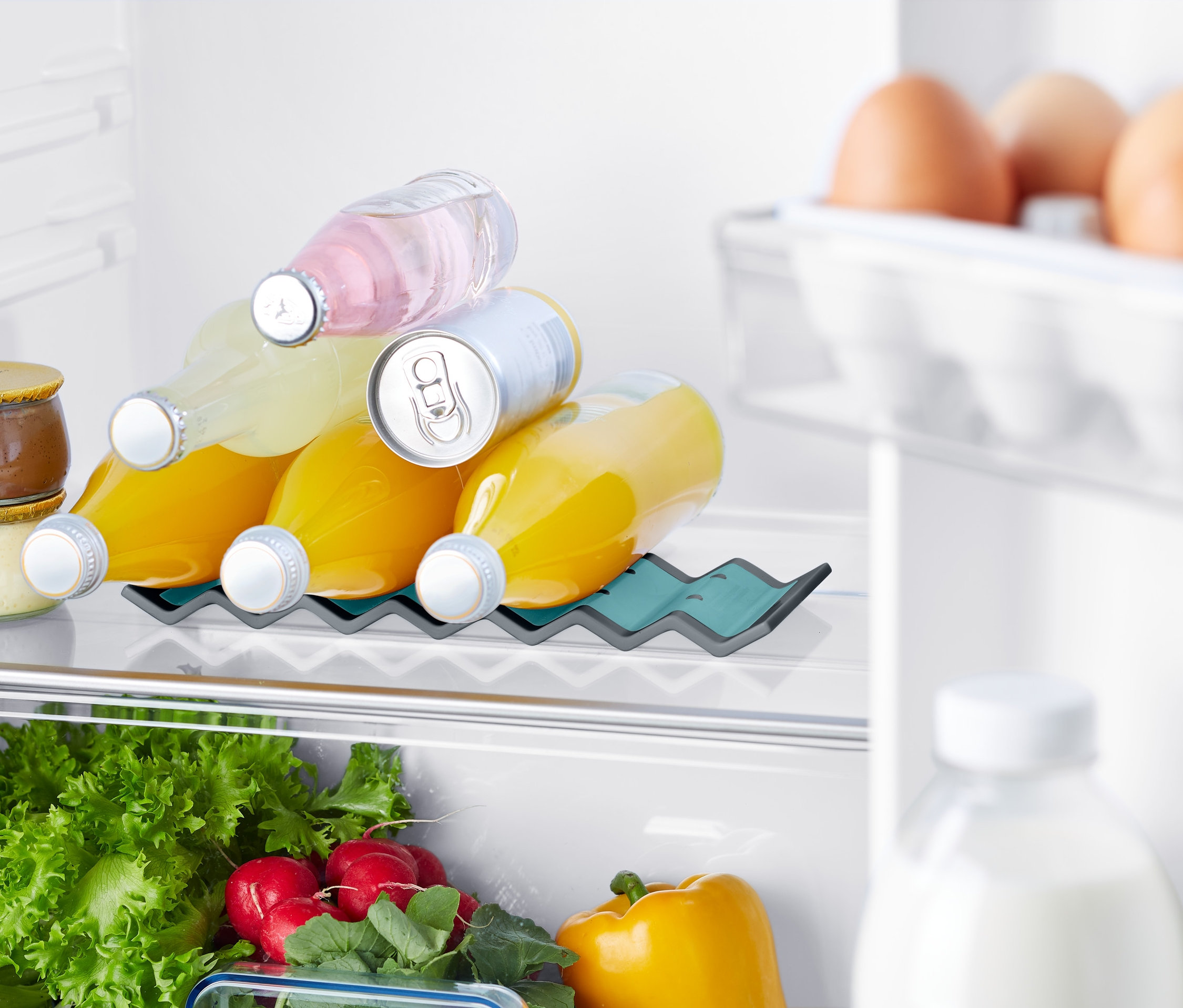 V chladničke je držiak na fľaše do chladničky, v ktorom sú umiestnené fľaše s nápojmi. Ďalej sú vidieť vajcia, zelenina a džemy.
