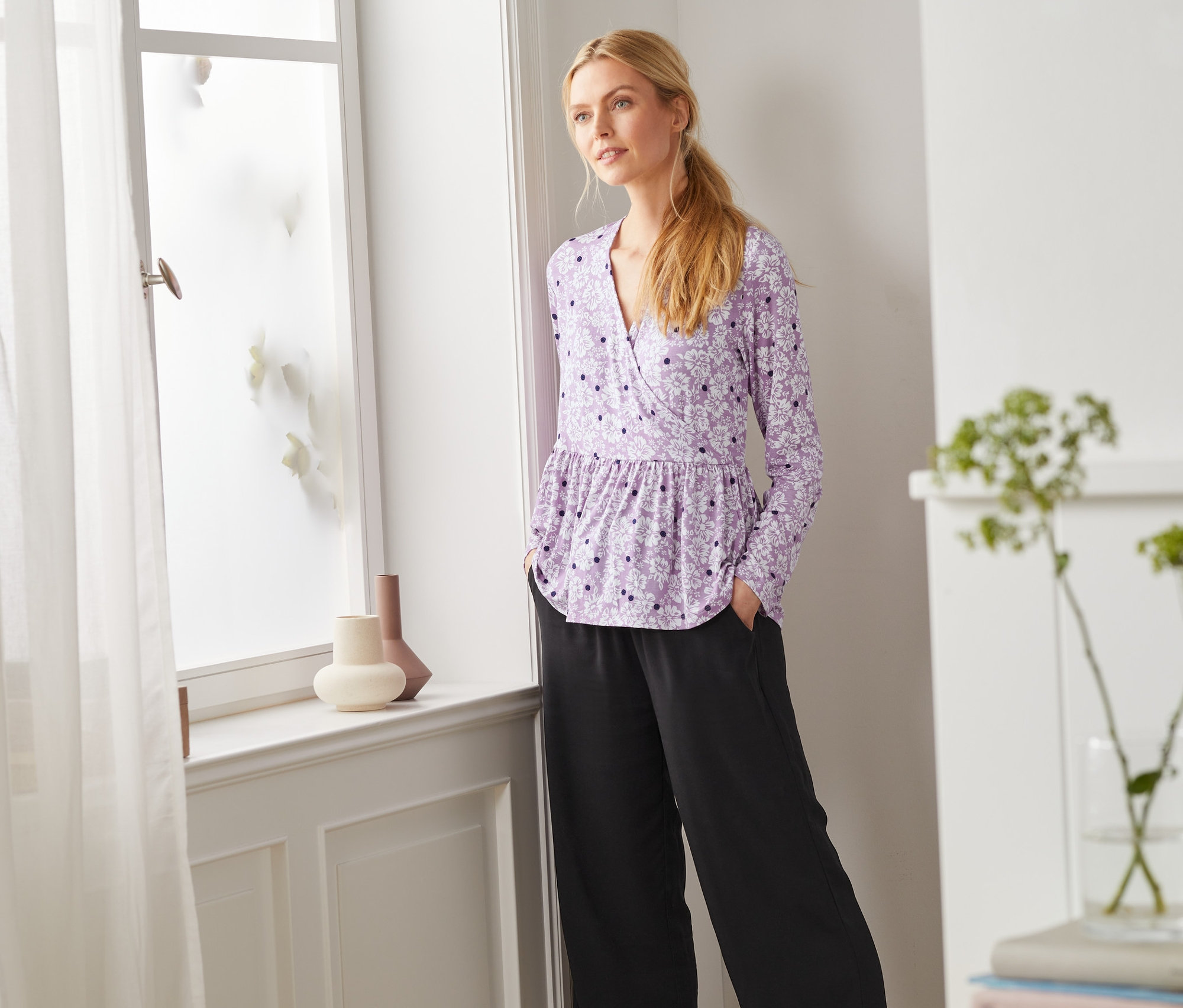 Žena stojí pri okne a má na sebe tričko s dlhým rukávom.