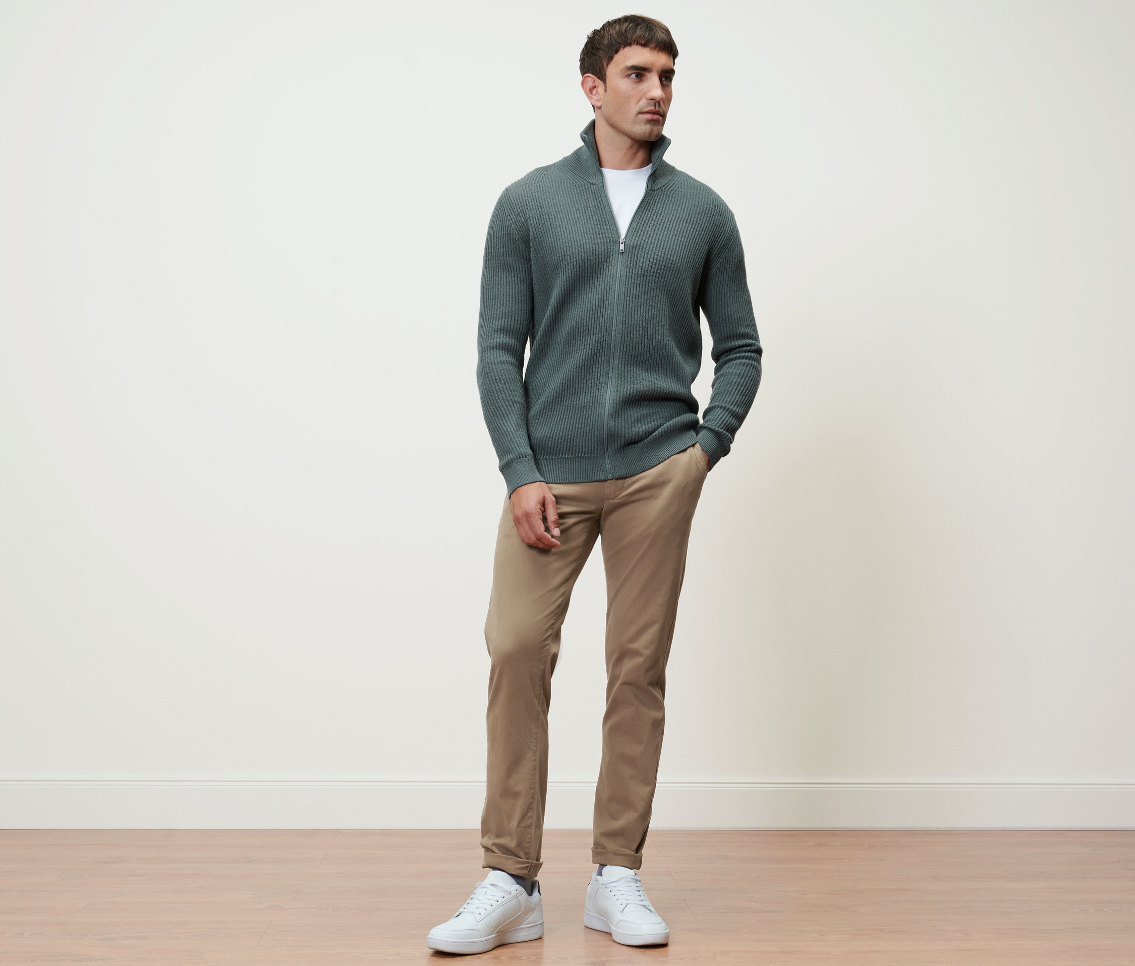 Muž stojí a má na sebe pletený sveter šalviovej farby a pieskové nohavice chino.