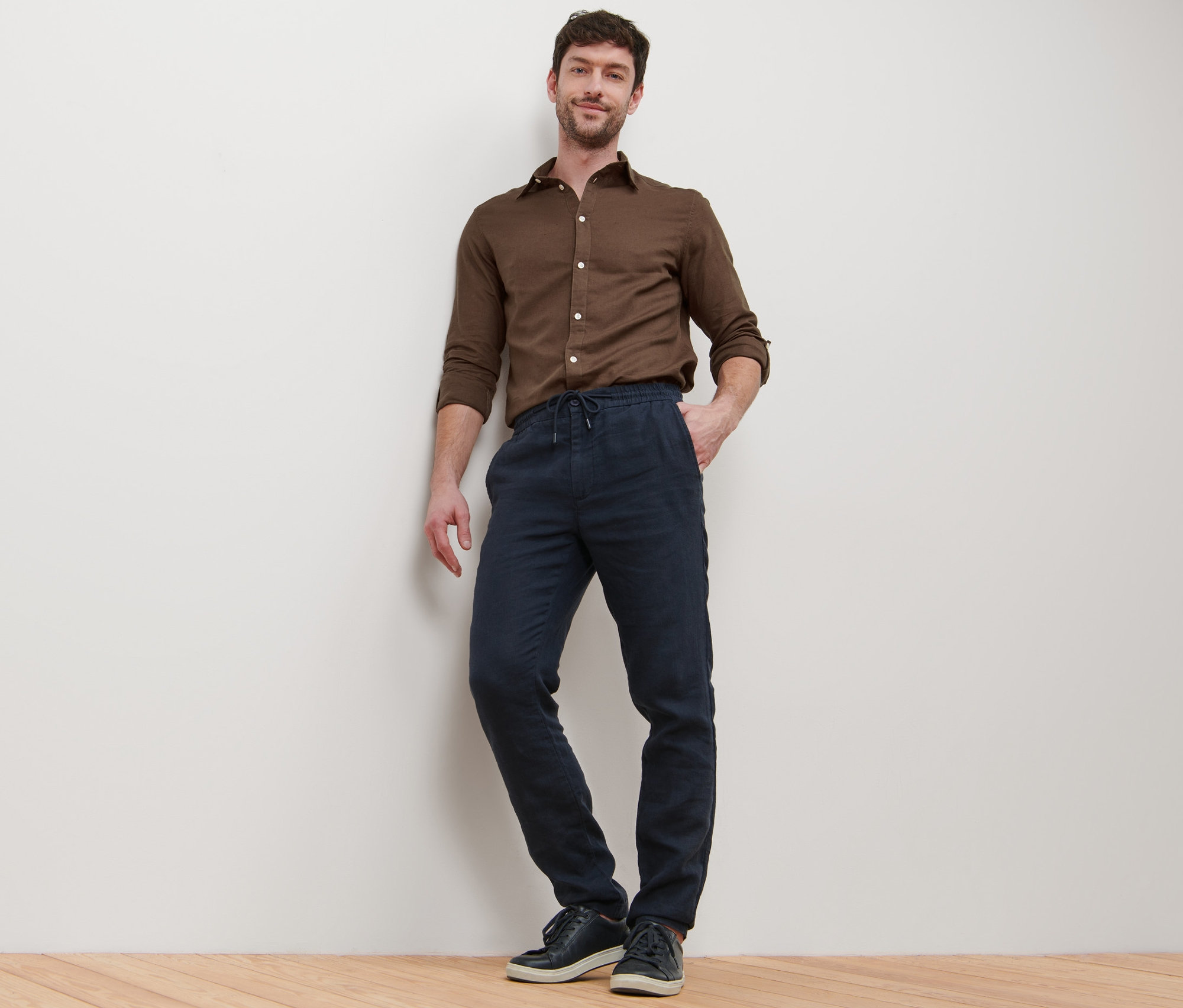 Muž opretý o stenu pózuje v hnedej košeli a ľanových nohaviciach chino.