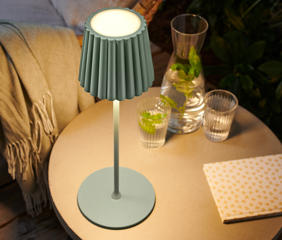 Na stole stojí zapnutá stolová LED lampa s akumulátorom v šalviovej farbe.