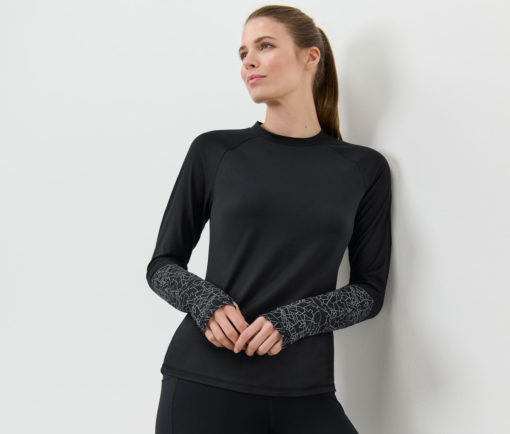 Žena opretá o stenu má na sebe čierne funkčné tričko s dlhými rukávmi v kombinácii čiernej a striebornej.