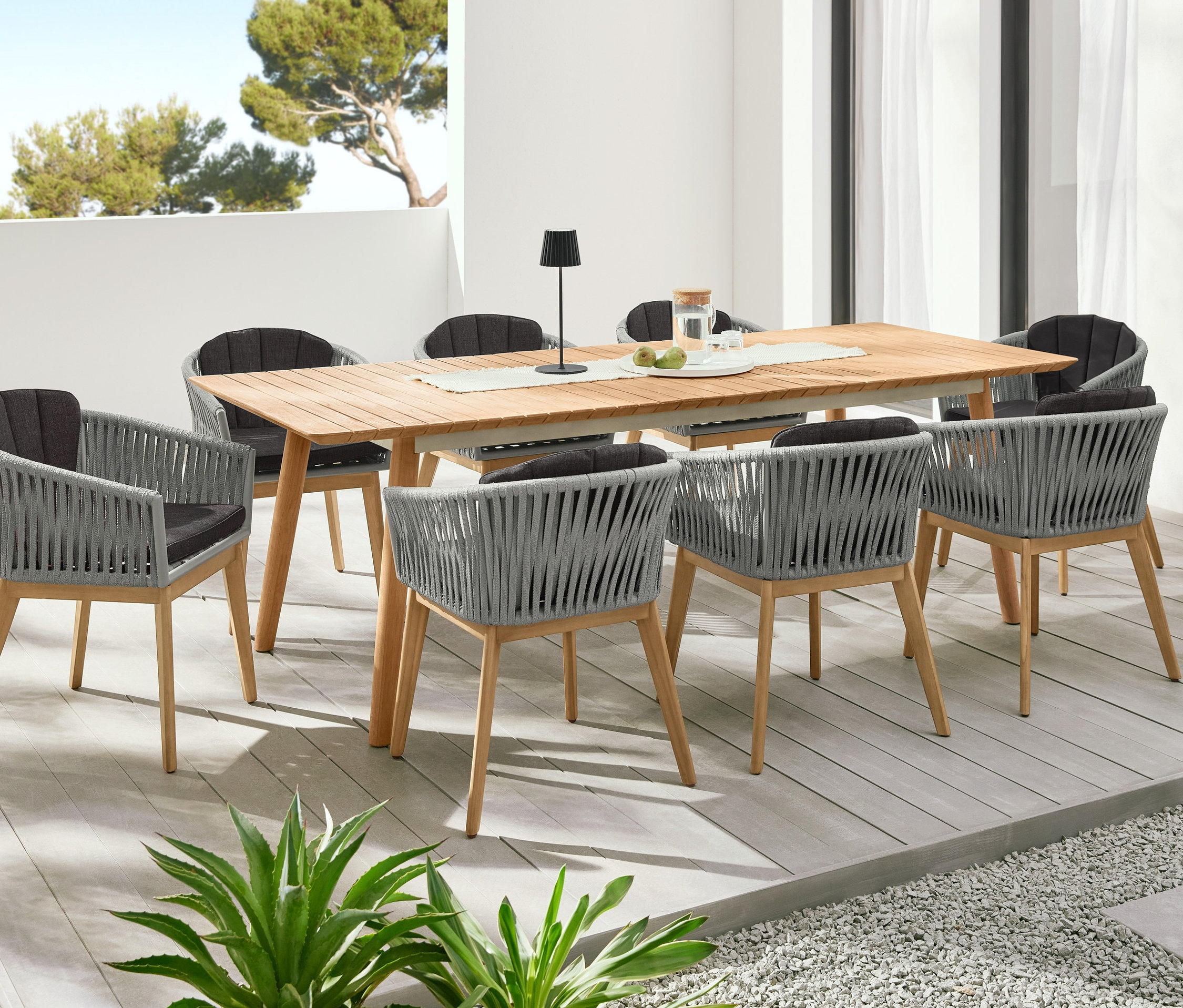 Teakový rozkladací stôl obklopený jedálenskými kreslami s textilným výpletom. V pozadí dva stĺpovité kvetináče »Tinus«, 2 ks.