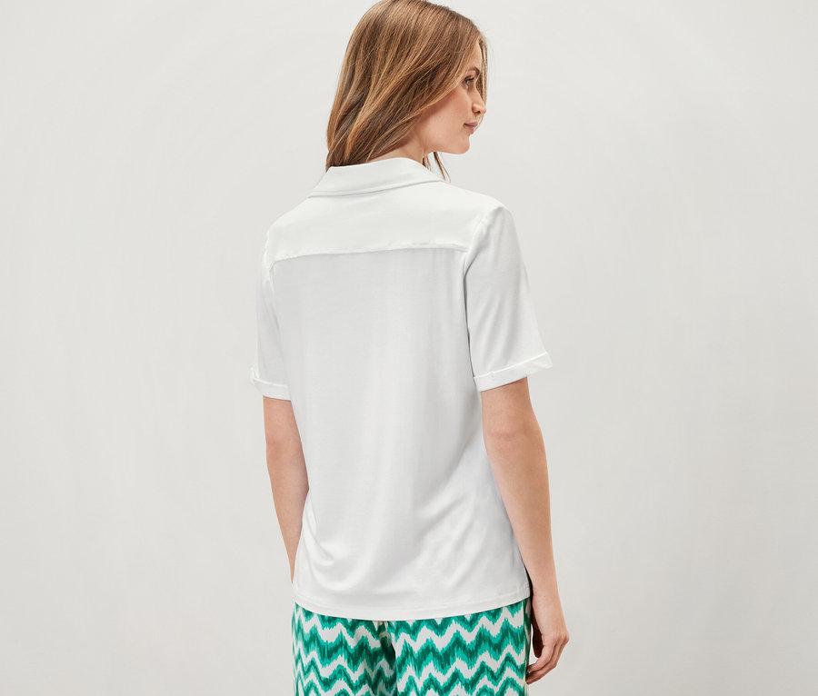 Žena stojí chrbtom k fotoaparátu a má na sebe biele blúzkové tričko s gombíkovou légou a zelené vzorované nohavice.