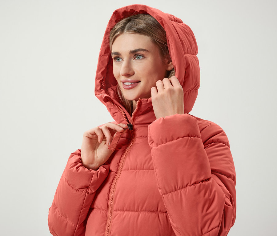 Žena si zapína zips na koralovočervenej páperovej bunde s kapucňou.