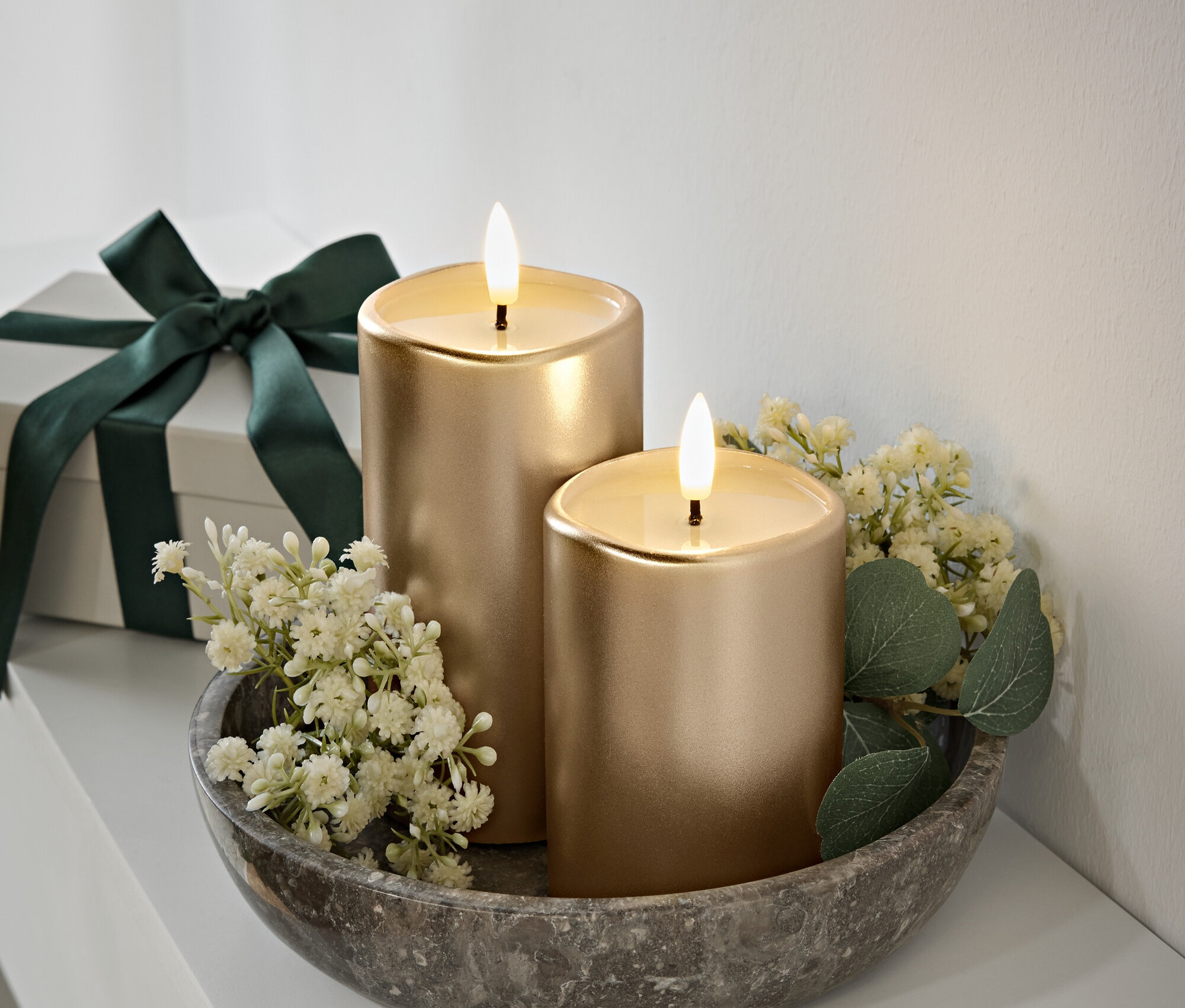 Dve zlaté sviečky s plameňmi v šedej kamennej miske, biele kvety a zelené listy. Darčeková krabička s tmavozelenou stuhou v pozadí.