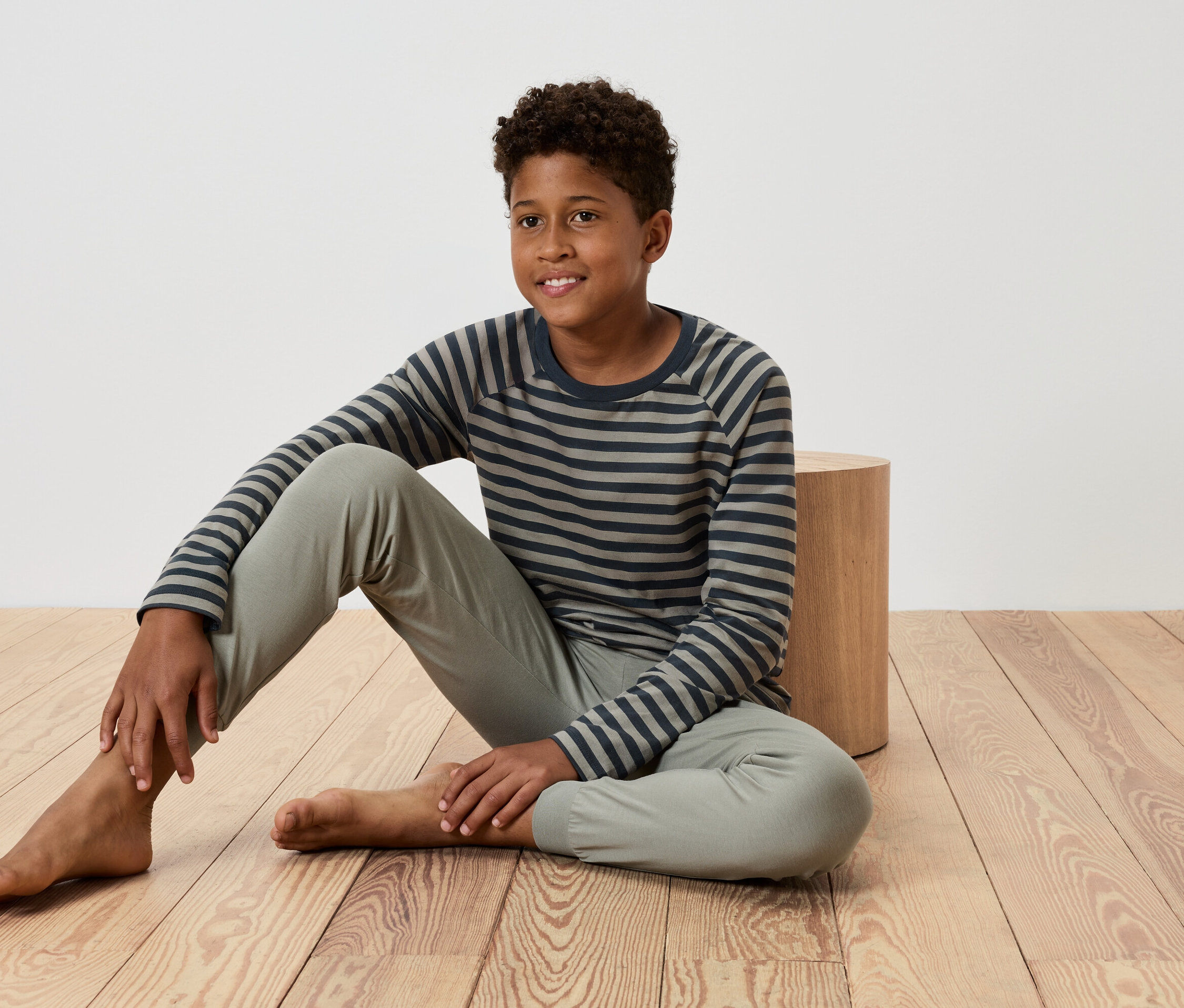 Mladý chlapec sedí na drevenej podlahe, opiera sa o malý drevený stolík, má na sebe pruhované tričko a nohavice.