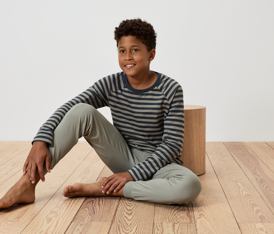 Mladý chlapec sedí na drevenej podlahe, opiera sa o malý drevený stolík, má na sebe pruhované tričko a nohavice.