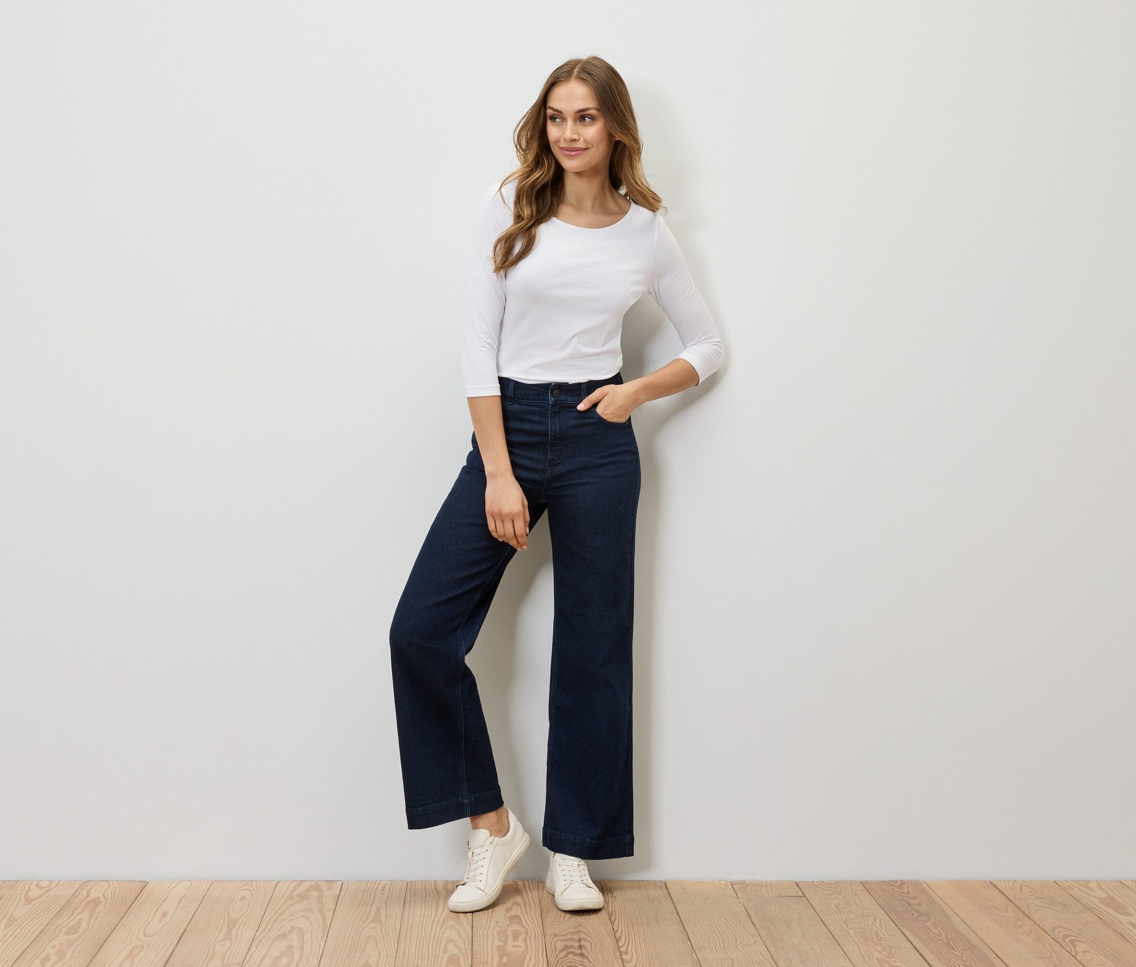 Žena pózuje v bielom tričku s trojštvrťovým rukávom, tmavomodrých širokých džínsoch a bielych teniskách.