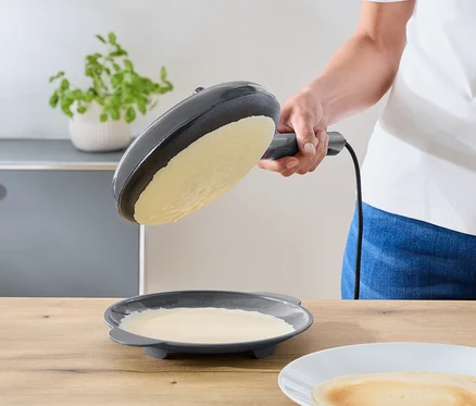 Osoba nakláňa tmavosivú elektrickú panvicu nad tanier so zvyškom cesta. Hotová palacinka leží na bielom tanieri na drevenej doske.