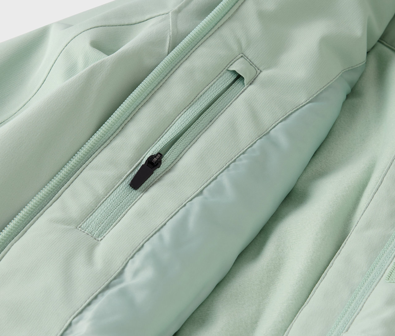 Detail svetlozelenej bundy s vreckom na zips a zipsom v strede.