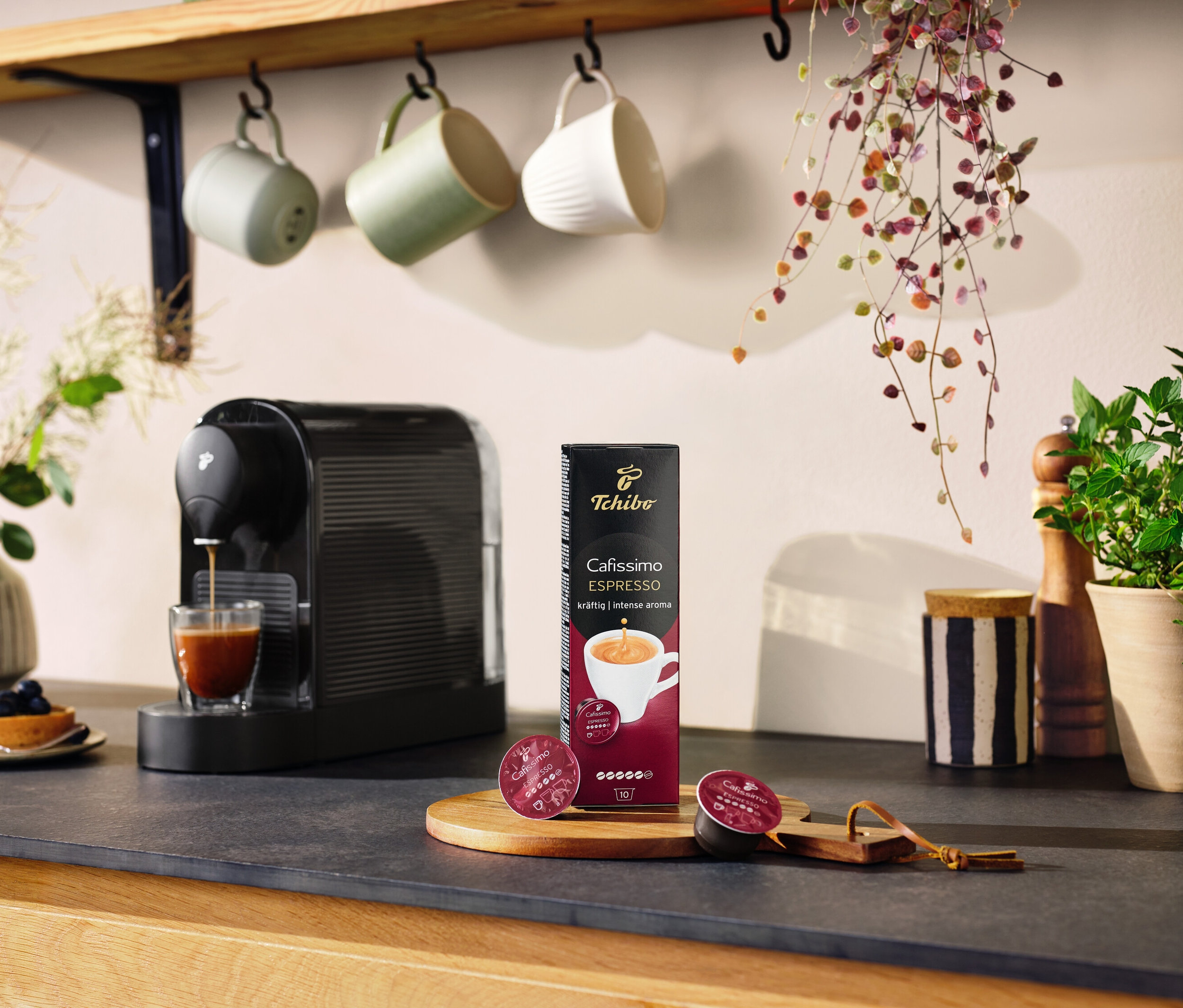 Čierny kávovar Cafissimo pure+ pripravuje kávu do sklenice. Na stole leží balenie kapsúl a dve kapsule.