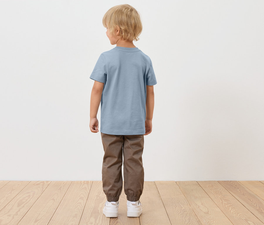 Chlapec s blond vlasmi stojí chrbtom, má na sebe modré tričko, hnedé nohavice a biele topánky.