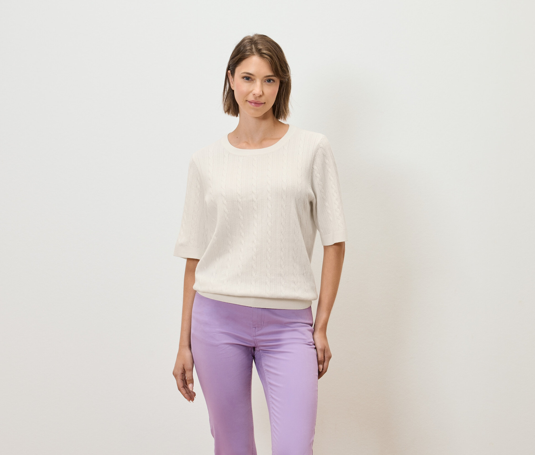 Žena pózuje na svetlom pozadí v krémovom svetri s krátkym rukávom a fialových nohaviciach.