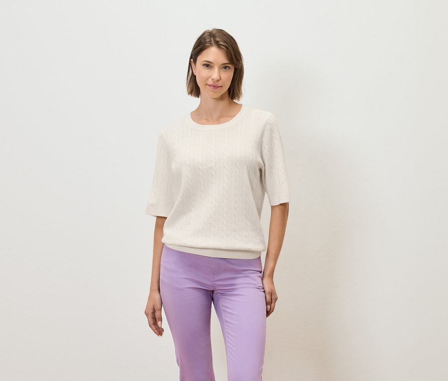 Žena pózuje na svetlom pozadí v krémovom svetri s krátkym rukávom a fialových nohaviciach.