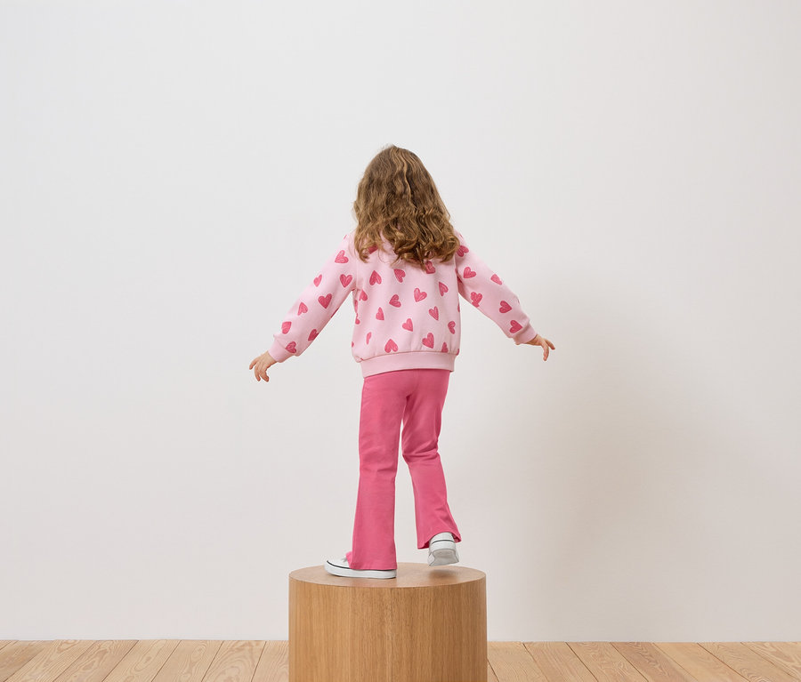 Dievča stojí chrbtom ku kamere na drevenom podstavci, má na sebe ružovú mikinu so srdiečkami a ružové nohavice.