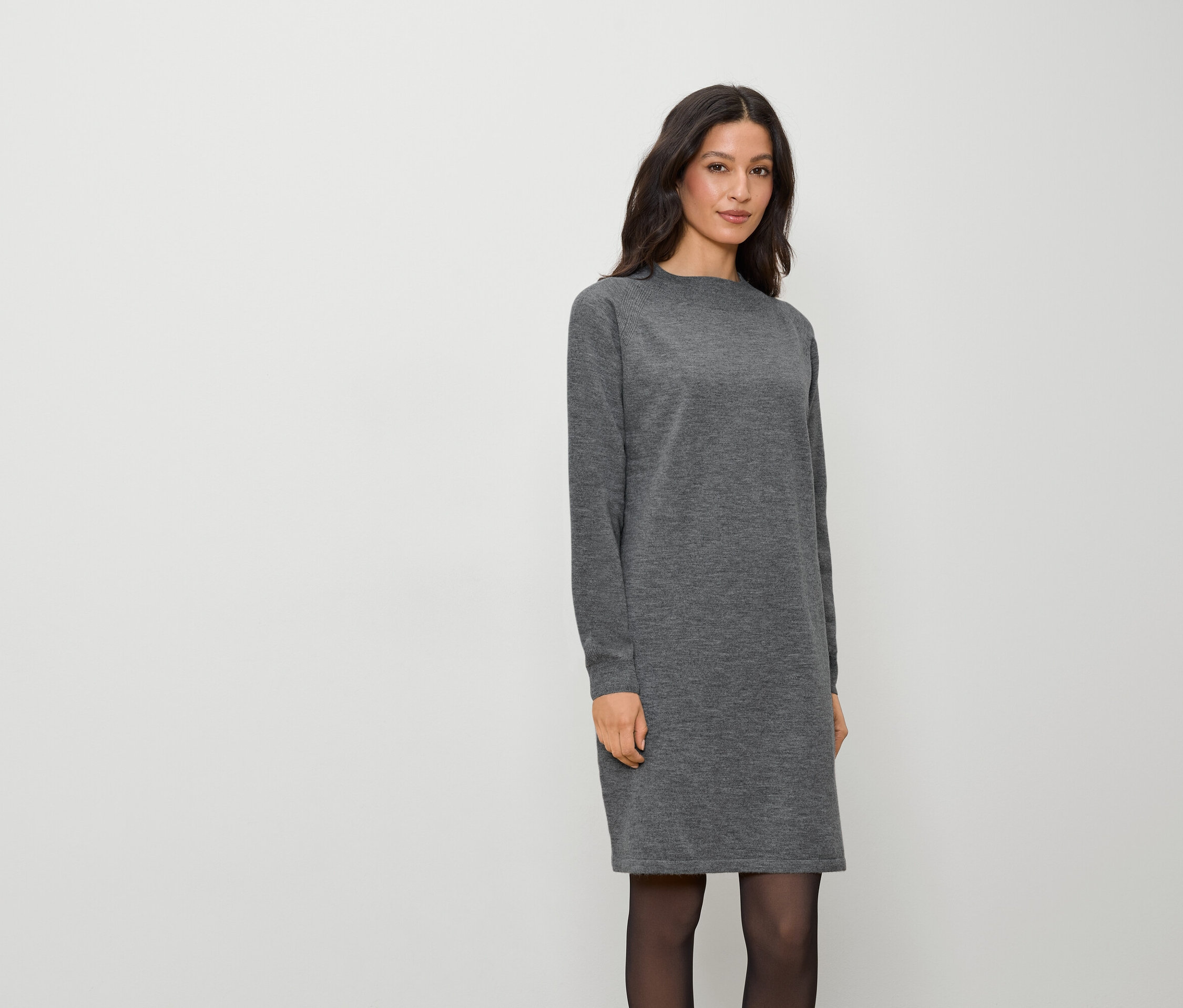 Žena v sivých šatách s dlhým rukávom a čiernych pančuchách stojí na bielom pozadí.