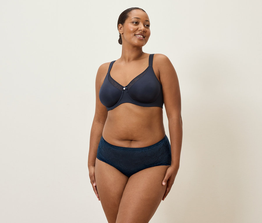 Žena stredného veku s tmavými vlasmi a tmavomodrou spodnou bielizňou stojí na krémovom pozadí.