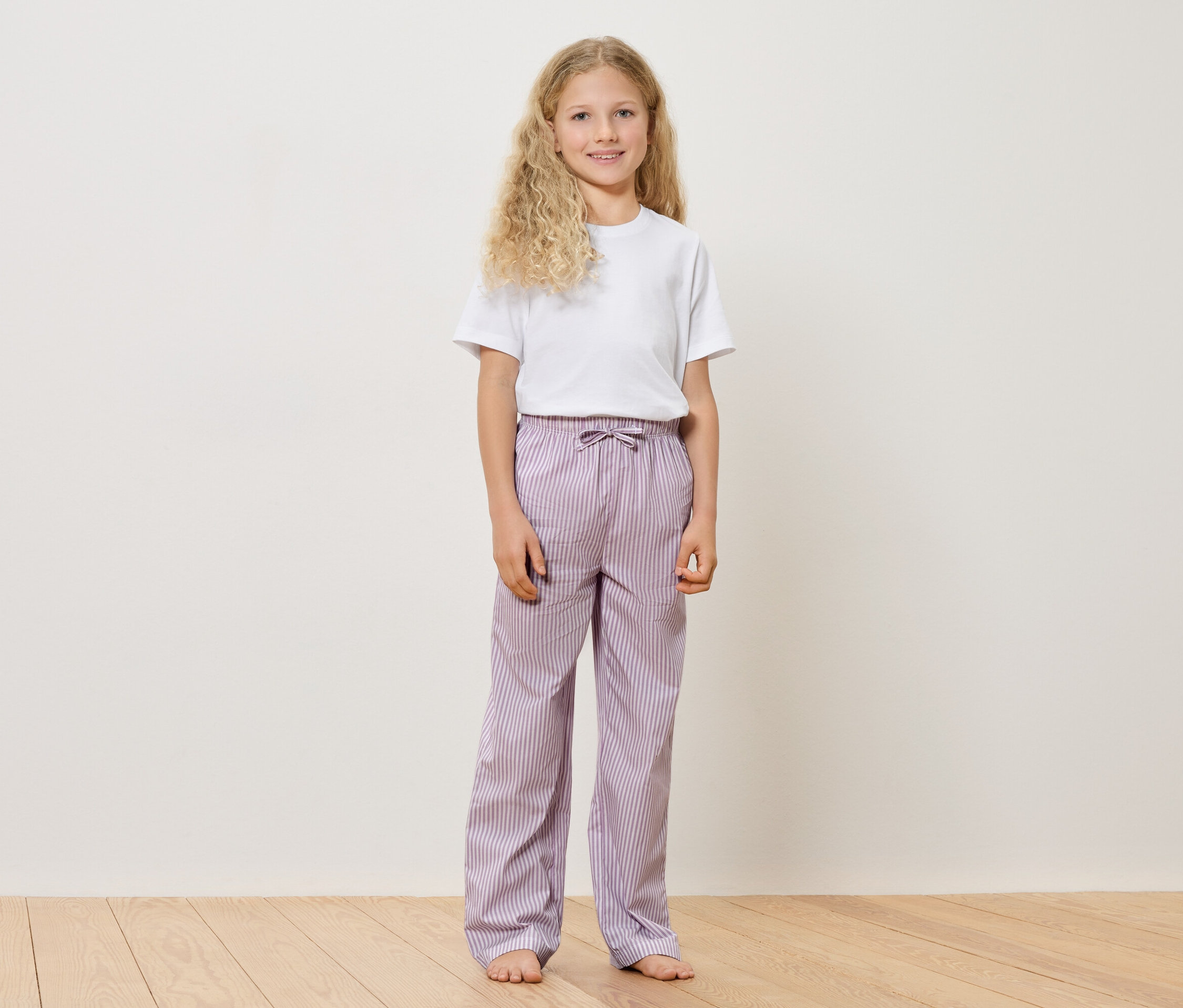 Dievča s blond, kučeravými vlasmi, v bielom tričku a fialových pruhovaných nohaviciach stojí bosé na drevenej podlahe pred bielou stenou.