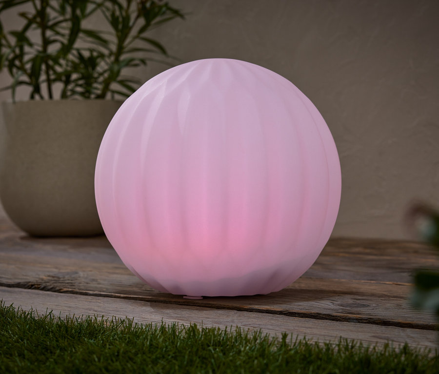 Na drevenom stole stojí ružová guľová lampa vedľa kvetináča s rastlinou.
