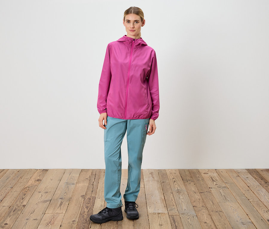 Žena stojí pred bielou stenou a má na sebe ružovú bundu, modré nohavice a čierne topánky.