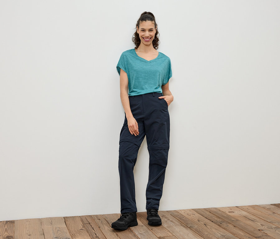 Žena stojí pred bielou stenou a má na sebe modrozelené tričko, tmavomodré nohavice so zipsom a čierne topánky.