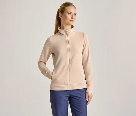 Žena s blond copom má na sebe béžovú rebrovanú bundu na zips a modré nohavice, pričom jednu ruku má vo vrecku bundy.