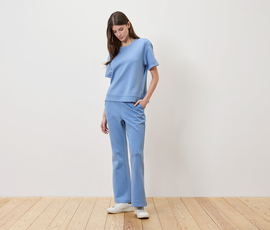 Žena s hnedými vlasmi má na sebe svetlomodré tričko a zodpovedajúce nohavice a ruku vo vrecku.