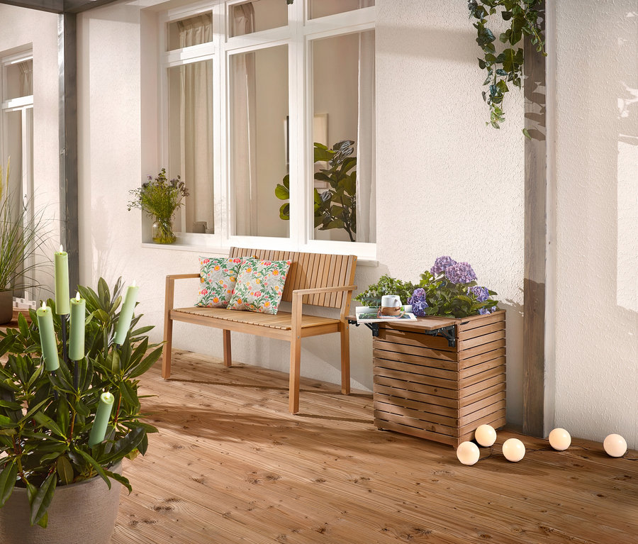 Na drevenej terase stojí drevená lavica s vankúšmi, kvetináč s kvetmi a črepníková rastlina so sviečkami.