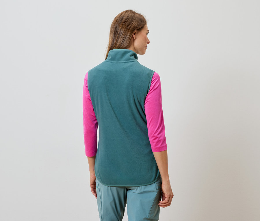 Pohľad zozadu na ženu s dlhými hnedými vlasmi v tyrkysovej fleecovej veste, ružovej košeli s dlhým rukávom a tyrkysových nohaviciach.