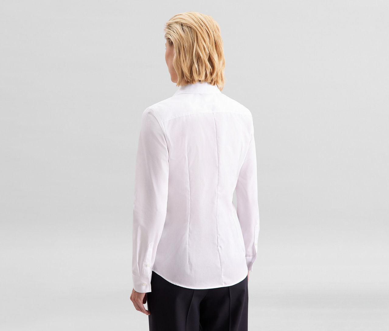 Žena zozadu, oblečená v bielej blúzke Seidensticker Slim Fit non iron a čiernych nohaviciach.