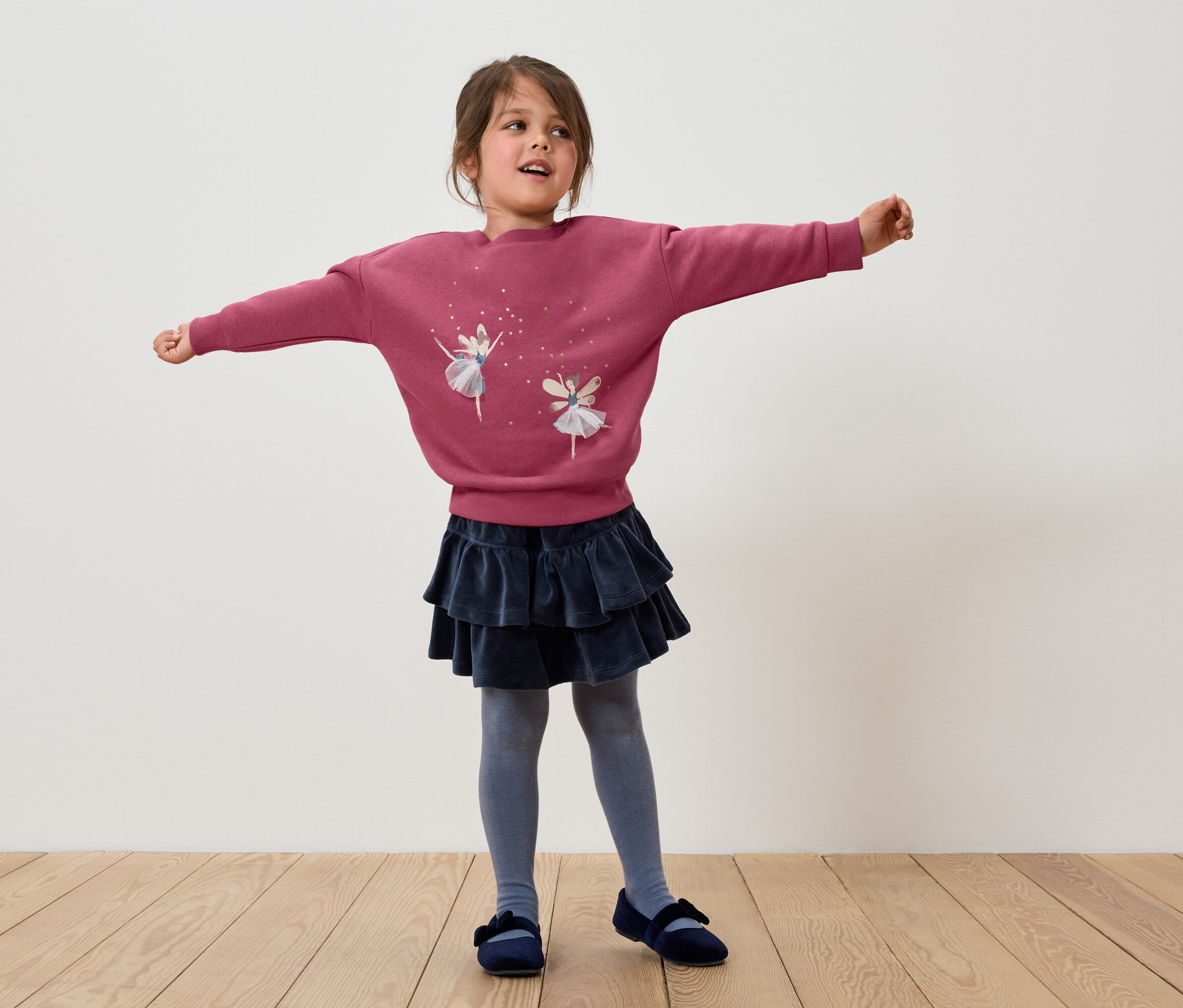 Dievča stojí s rozpaženými rukami, má na sebe ružovú mikinu s baletkami, tmavomodrú sukňu s volánmi, sivé pančuchy a modré topánky.