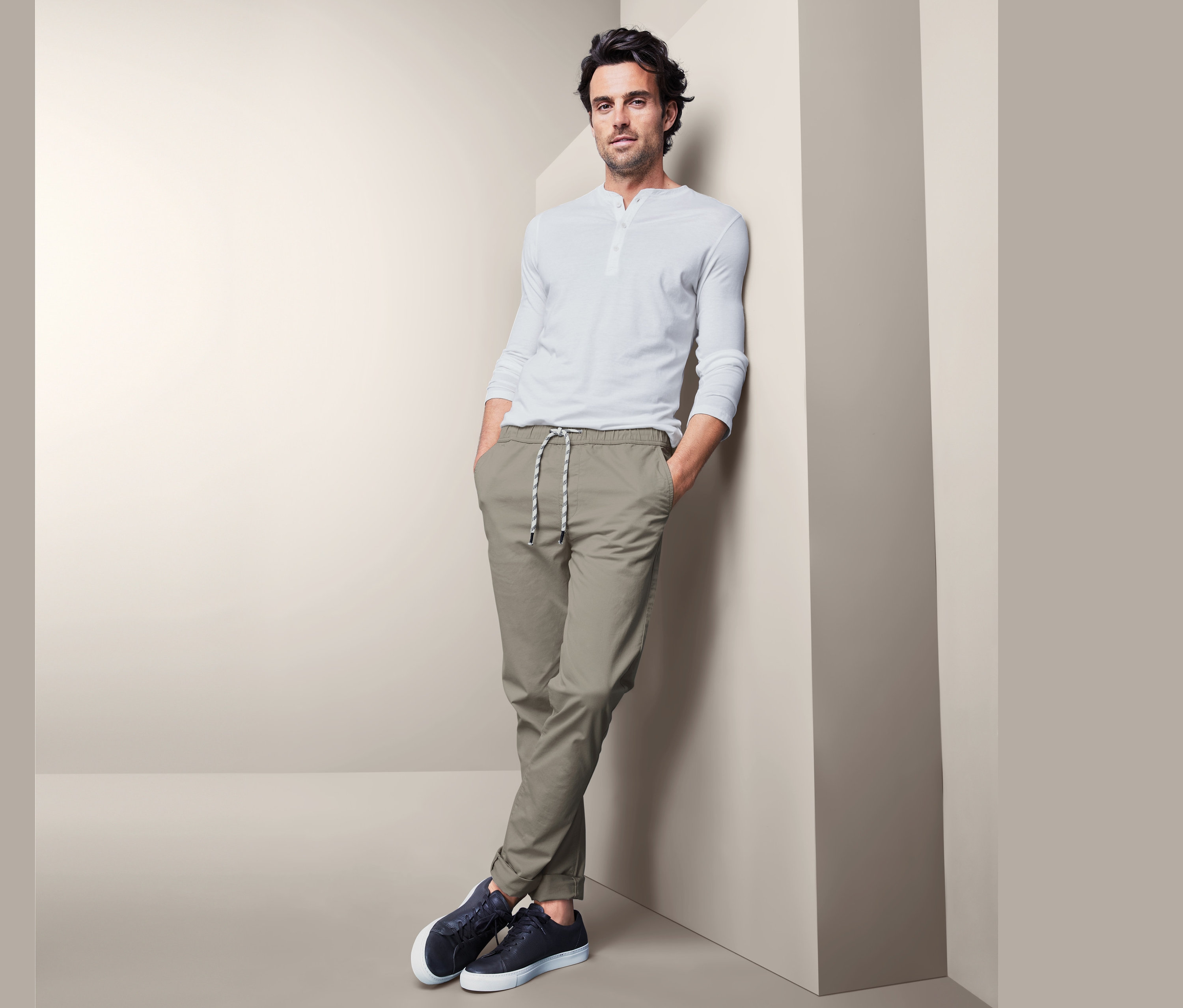 Muž opretý o stenu má na sebe pohodlné chino nohavice, biele tričko a tmavomodré tenisky.